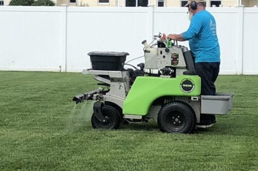 Lawn care technician operating a ride-on spreader-sprayer applying fertilizer or treatment to a residential back yard.