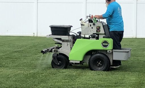 Lawn care technician operating a ride-on spreader-sprayer applying fertilizer or treatment to a residential back yard.