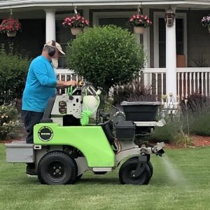 Lawn care technician operating a ride-on spreader-sprayer applying fertilizer or treatment to a residential front lawn.