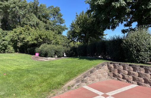 Commercial property grounds maintenance with mowed lawn, trimmed shrub borders, stone retaining wall, and brick paver patio area.