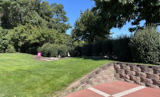 Commercial property grounds maintenance with mowed lawn, trimmed shrub borders, stone retaining wall, and brick paver patio area.